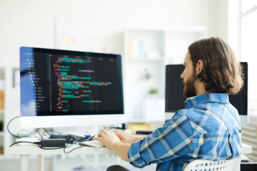 Serious pensive hipster young bearded coder in casual shirt creating computer software and working with desktop computer in modern office